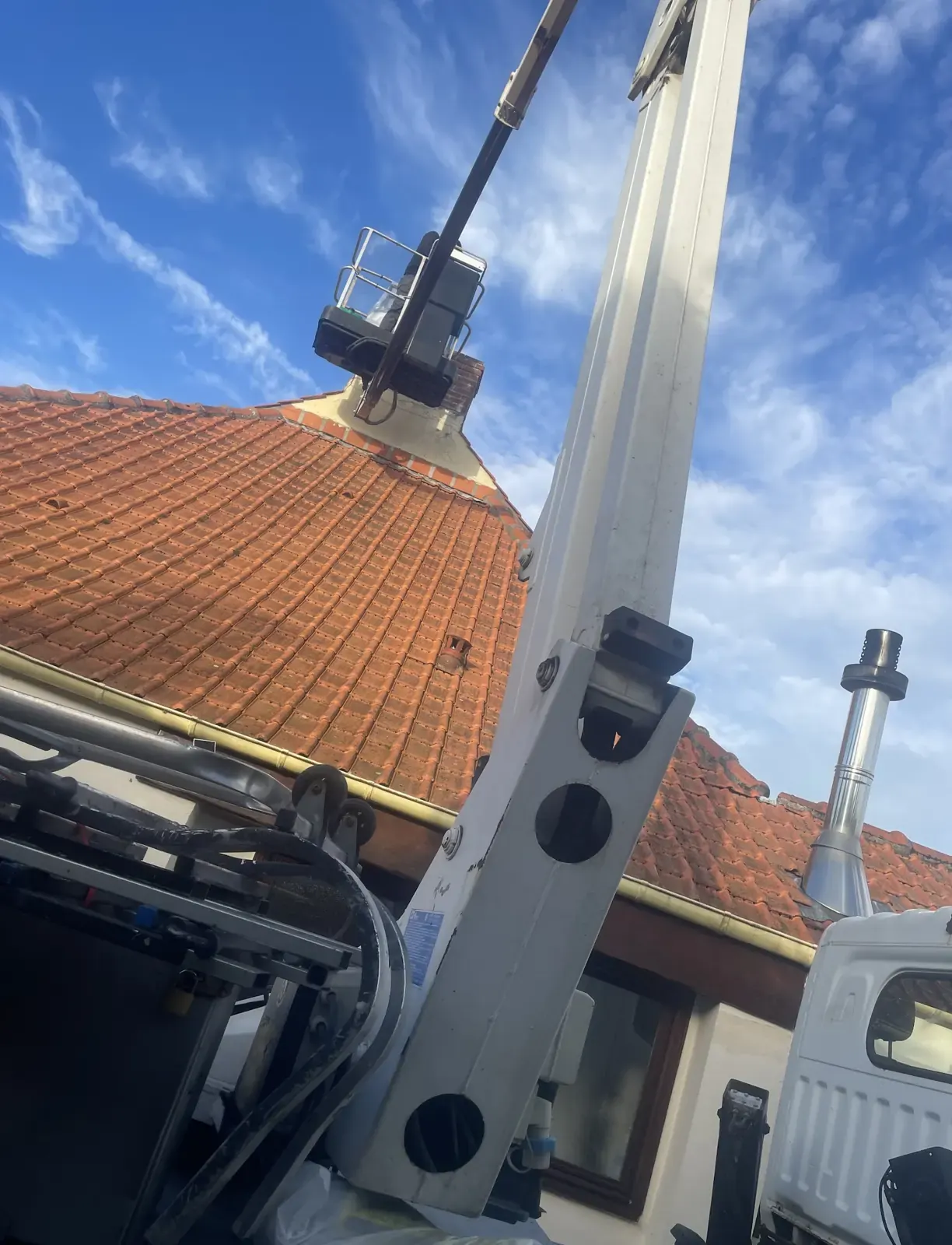 Bras de la nacelle déployé le long d’une toiture en tuiles orangées, ciel bleu